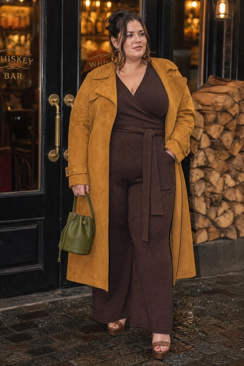 A woman wears a mustard long coat over a dark brown wrap-style wide-leg jumpsuit with an olive green drawstring bag and platform sandals.