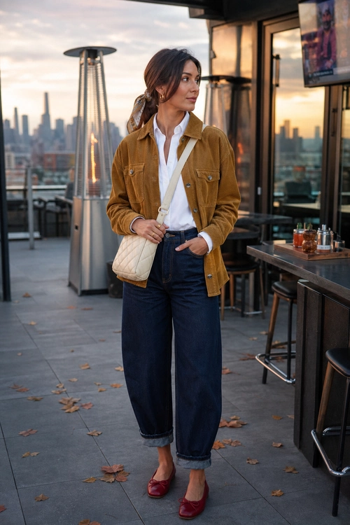Woman wearing a mustard jacket, white button-up shirt, dark cuffed wide-leg jeans, burgundy flats, and a cream quilted crossbody bag.