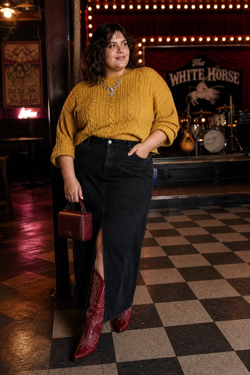 A woman wears a mustard cable-knit sweater with a black front-slit denim skirt, burgundy western boots, a matching mini handbag, and layered necklaces.