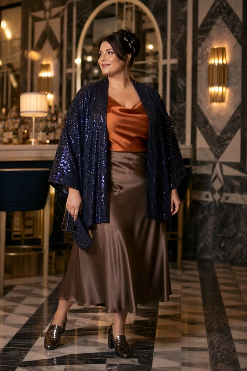 A woman wears a navy sequin kimono jacket over a rust satin top with a chocolate brown satin midi skirt and dark loafers.