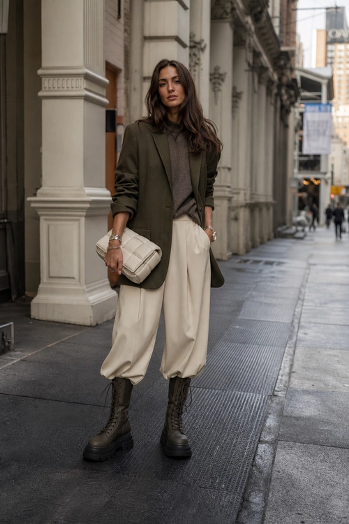 Olive Blazer and Cream Trousers A woman wears an olive blazer, taupe knit top, cream trousers, dark lace-up boots, and carries a quilted cream bag on a city sidewalk.