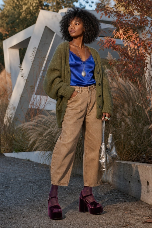 A woman wears an olive green cardigan over a blue satin camisole with tan cropped trousers, burgundy platform heels, and a metallic silver handbag.