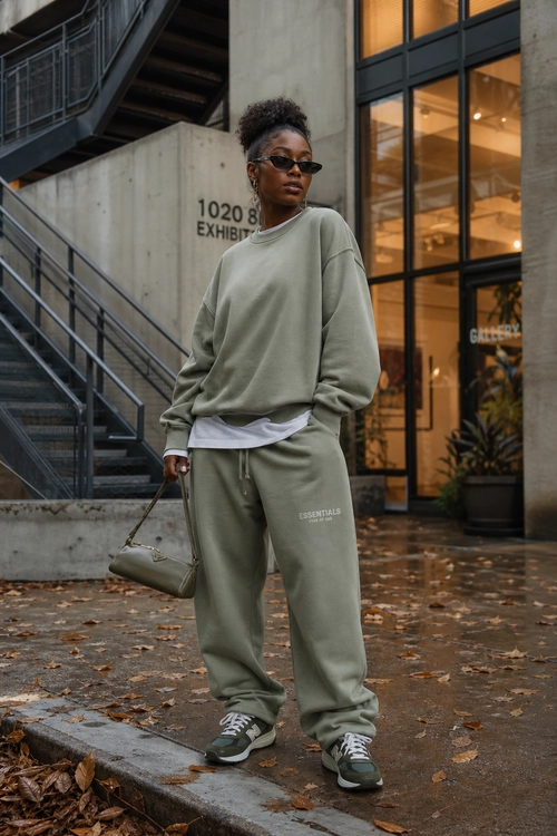 A person wears an oversized olive green sweatshirt with matching joggers, a white layered T-shirt, sunglasses, sneakers, and a taupe handbag.