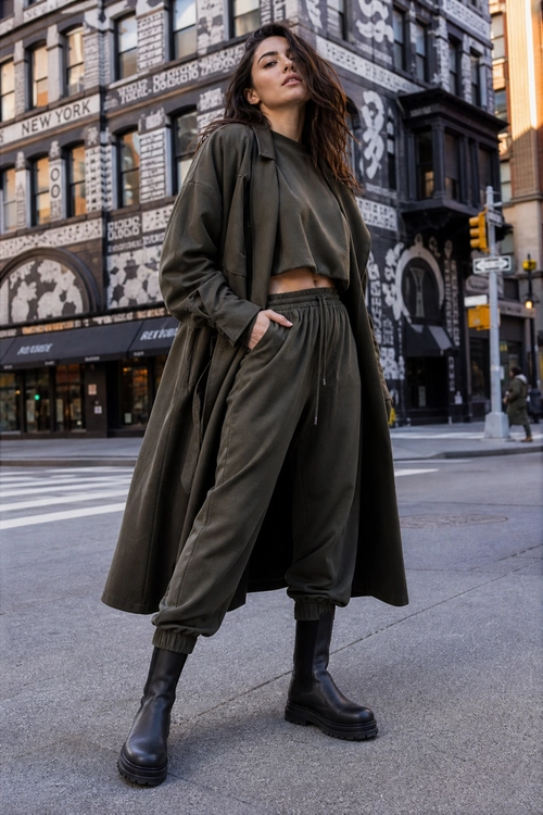 Olive Layered Streetwear Look A model wears a dark olive long coat over a cropped top and matching jogger pants with black lug-sole boots on a city street.