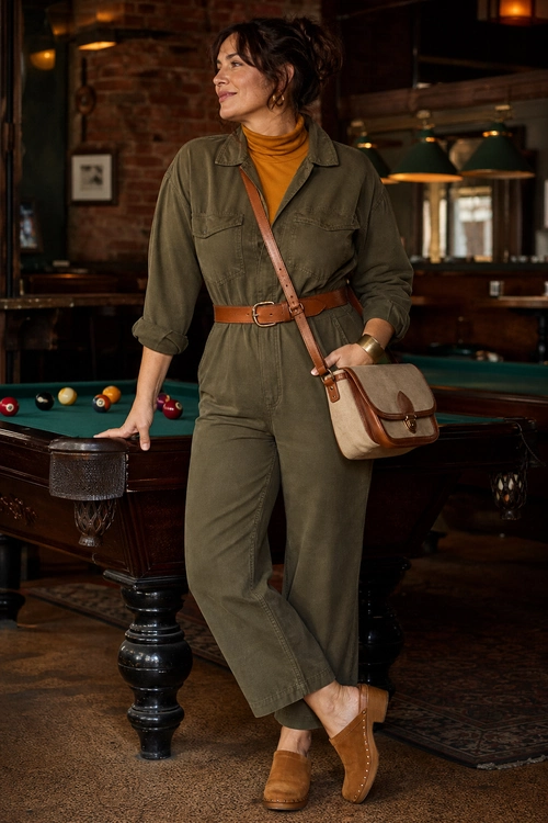 A woman wears an olive utility jumpsuit over a mustard turtleneck with a brown belt, crossbody bag, and tan clogs.