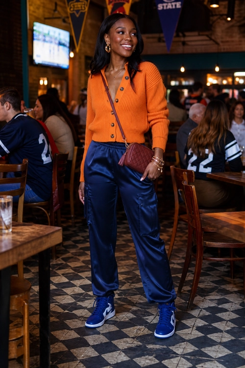 Orange Cardigan and Navy Joggers A woman wears an orange button-front cardigan with navy satin jogger pants, blue high-top sneakers, and a burgundy crossbody bag.