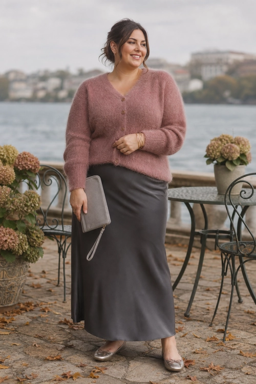 A woman wears a soft pink button-front cardigan with a gray satin skirt, metallic flats, gold bracelets, and a gray wristlet clutch.
