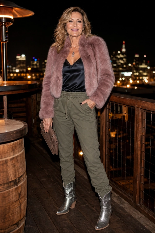A woman wears a pink plush jacket over a black satin camisole with olive drawstring pants, metallic ankle boots, a taupe clutch, and a pendant necklace.