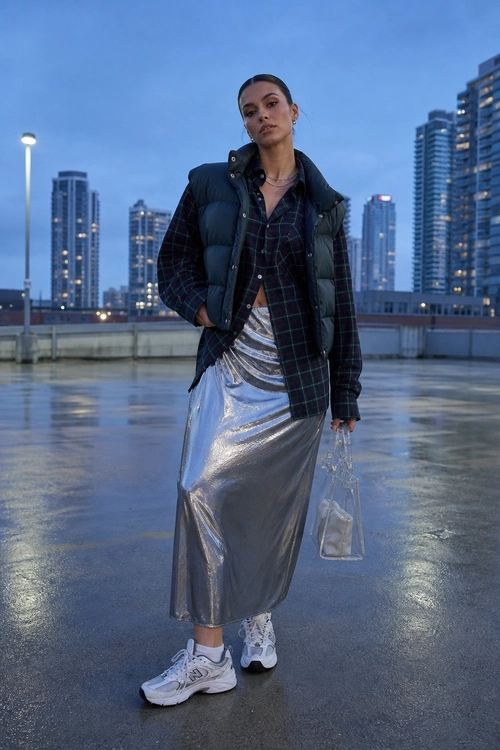 Plaid Shirt with Metallic Skirt A layered outfit featuring a dark plaid shirt, black puffer vest, metallic silver midi skirt, white sneakers, and a clear handbag.