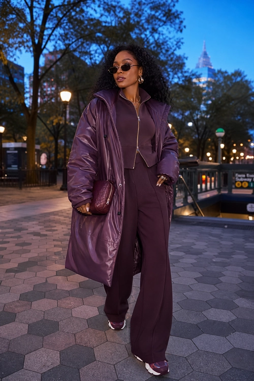 A woman wears a glossy plum long coat over a matching fitted top and wide-leg trousers, styled with sunglasses, a shoulder bag, and sneakers.