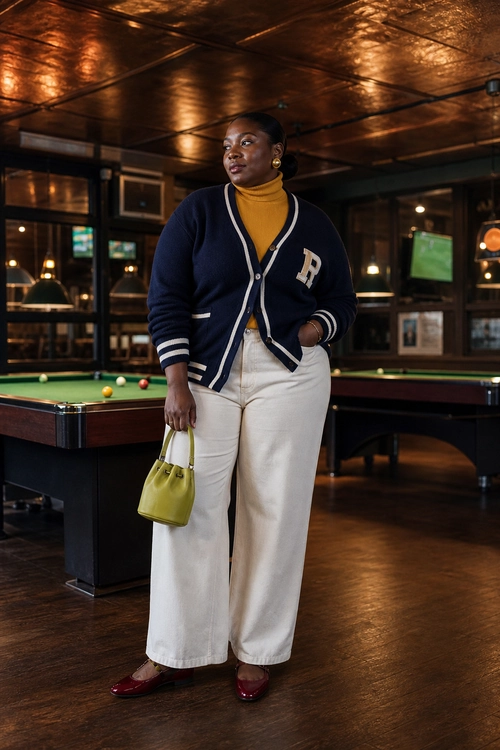 Preppy Cardigan Outfit A woman wears a navy varsity-style cardigan over a mustard turtleneck with cream wide-leg pants, burgundy flats, and an olive green handbag.