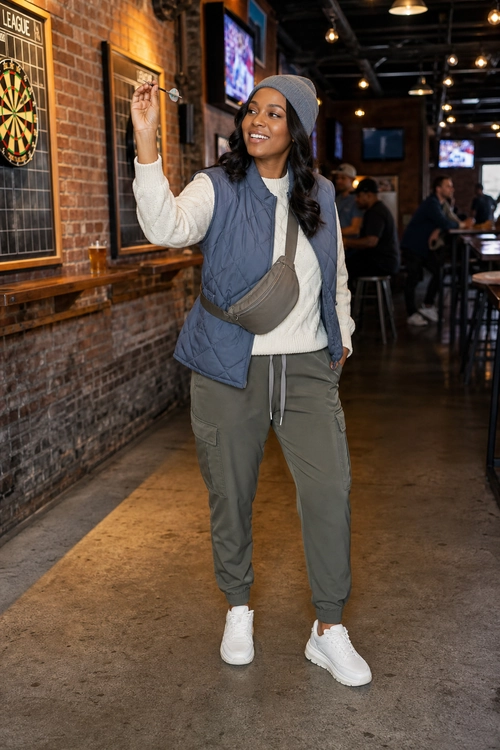 Casual Quilted Vest Outfit A woman wears a gray beanie, blue quilted vest, cream sweater, olive cargo joggers, crossbody bag, and white sneakers while playing darts.
