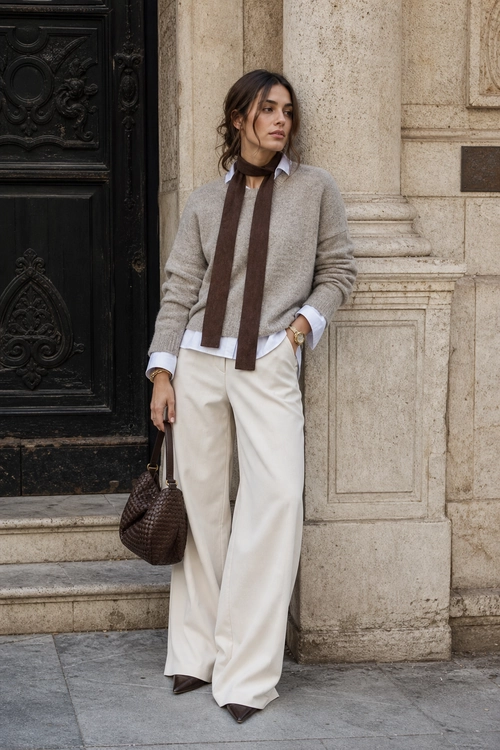 Refined Layered Neutral Outfit A woman wears a gray sweater over a white collared shirt with a brown scarf, wide-leg cream trousers, brown pointed shoes, and a woven brown handbag.