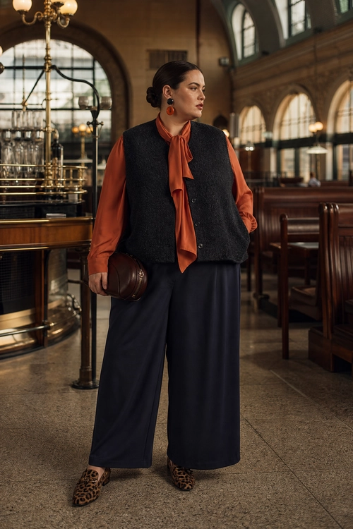 Rust Blouse and Wide-Leg Trousers A woman wears a rust tie-neck blouse with a dark sleeveless vest, wide-leg trousers, leopard print shoes, a brown clutch, and drop earrings.