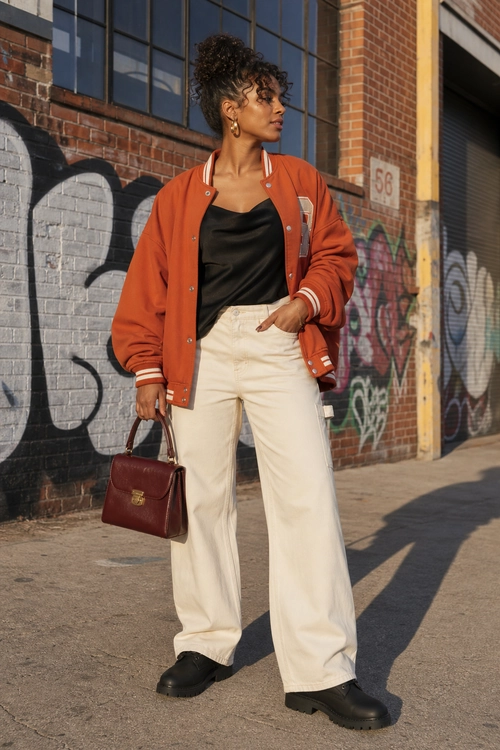 Rust Bomber Jacket and Wide-Leg Trousers A person wears a rust orange bomber jacket over a black top with cream wide-leg trousers, black shoes, and a burgundy handbag.