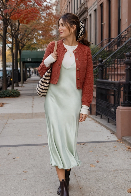 Woman wearing a rust cardigan over a pale mint slip dress with dark ankle boots and a striped shoulder bag on a city sidewalk.