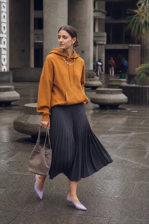 Rust Hoodie with Pleated Midi Skirt A rust-orange hoodie is paired with a dark pleated midi skirt, lavender pointed heels, a taupe tote bag, and a layered necklace.