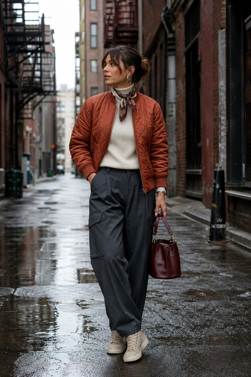 An urban outfit featuring a rust quilted jacket, cream turtleneck, patterned neck scarf, dark gray wide-leg trousers, beige sneakers, and a burgundy tote bag.