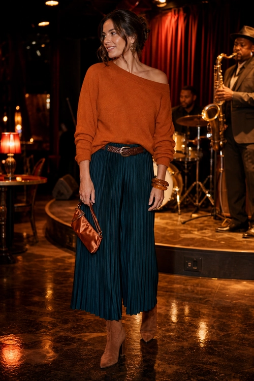 Rust Sweater and Pleated Skirt A woman wears a rust off-the-shoulder sweater with a teal pleated midi skirt, brown belt, clutch, bracelets, and ankle boots.