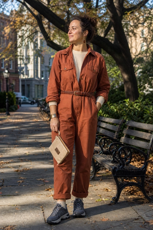 Rust Utility Jumpsuit Outfit A rust utility jumpsuit layered over a cream sweater and styled with gray sneakers and a beige clutch on a tree-lined sidewalk.