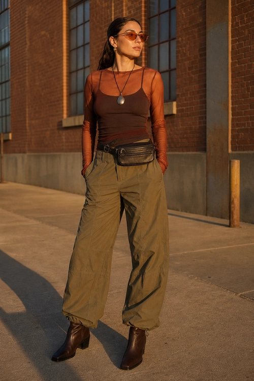 A fashion outfit featuring a sheer rust-brown long-sleeve top layered over a dark brown camisole, olive wide-leg trousers, a waist bag, sunglasses, and brown ankle boots.