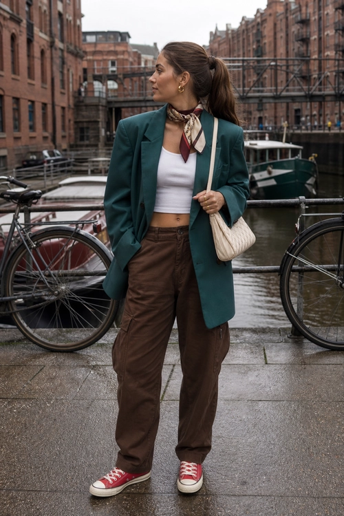 A city outfit featuring a teal blazer, white cropped top, brown trousers, red canvas sneakers, a patterned neck scarf, and a cream shoulder bag.