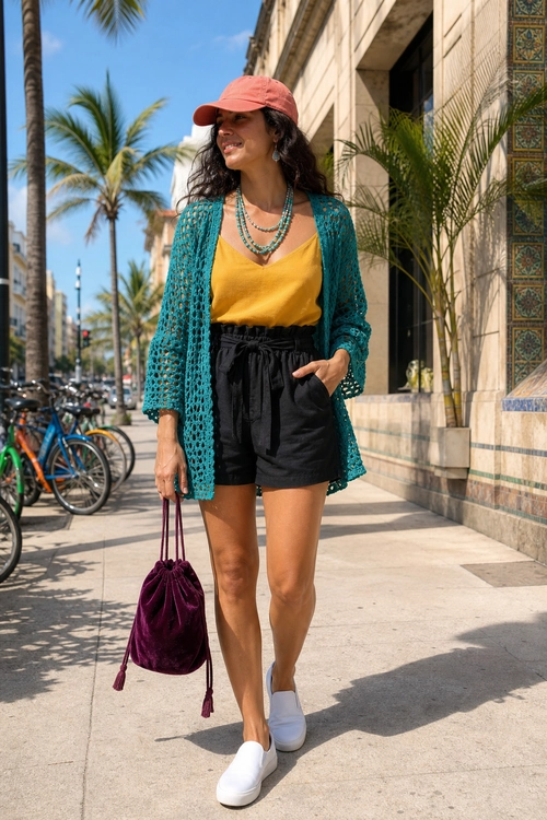 A woman wears a teal crochet cardigan over a mustard camisole with black shorts, white slip-on sneakers, a coral cap, layered necklaces, and a burgundy drawstring bag.