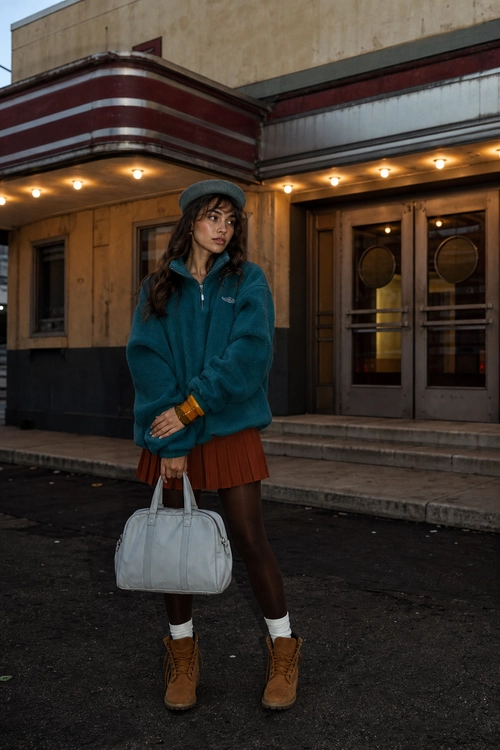 Teal Fleece and Pleated Skirt Outfit A teal quarter-zip fleece pullover styled with a rust pleated mini skirt, dark tights, brown ankle boots, a gray newsboy cap, and a light gray handbag.