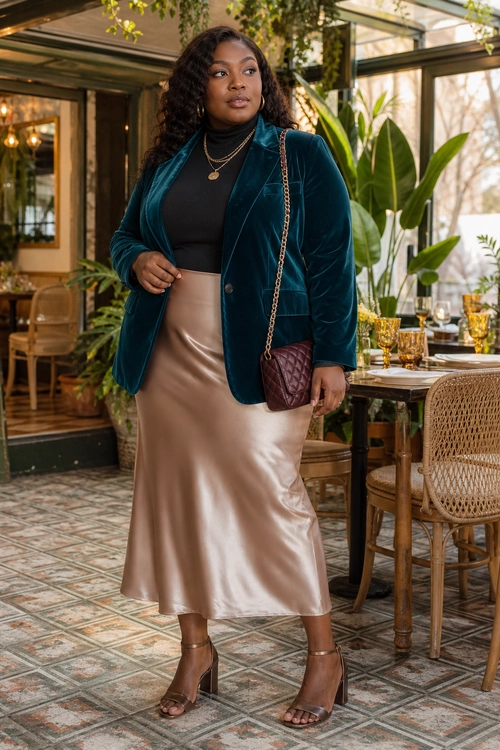 A woman wears a teal velvet blazer over a black top with a champagne satin midi skirt, heeled sandals, and a burgundy chain-strap bag.