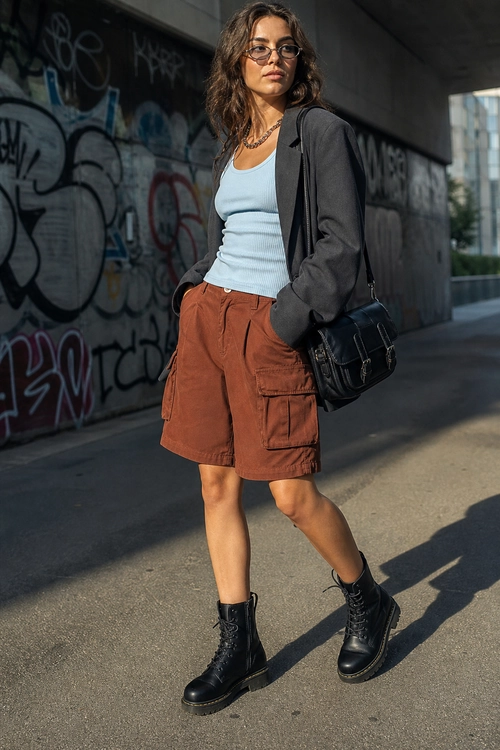 Urban Blazer and Cargo Shorts Outfit Woman wearing a charcoal blazer over a light blue ribbed top with rust cargo shorts, black combat boots, glasses, and a shoulder bag.