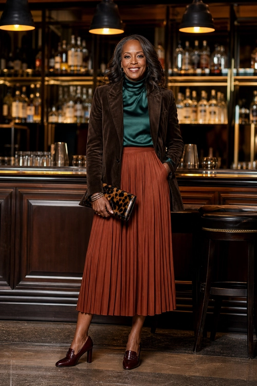 Velvet Blazer and Pleated Skirt A woman wears a dark brown velvet blazer with a teal turtleneck top, rust pleated midi skirt, leopard clutch, and burgundy heeled loafers.
