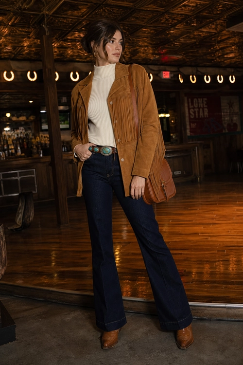 A woman wears a tan fringe jacket over a cream turtleneck with dark flare jeans, a western belt, brown boots, and a brown shoulder bag.