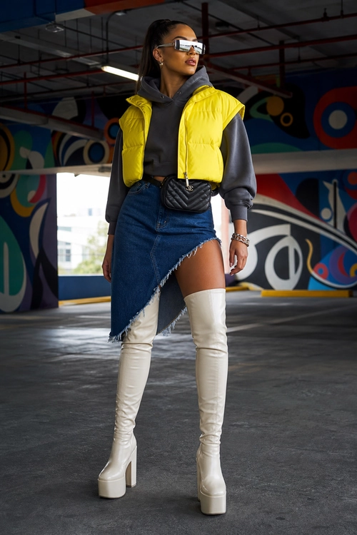 Yellow Puffer Vest and Denim Skirt Outfit A fashion outfit featuring a yellow quilted puffer vest over a charcoal hooded sweatshirt, an asymmetrical denim skirt, and white thigh-high platform boots.