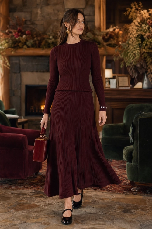 Burgundy Ribbed Knit Skirt Set Woman wearing a burgundy ribbed knit top and flowing pleated skirt with black Mary Jane shoes and a structured handbag.