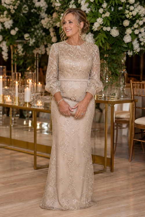 Champagne Embroidered Evening Gown A woman wears a champagne floor-length gown with long sleeves, floral embroidery, and a fitted waist.