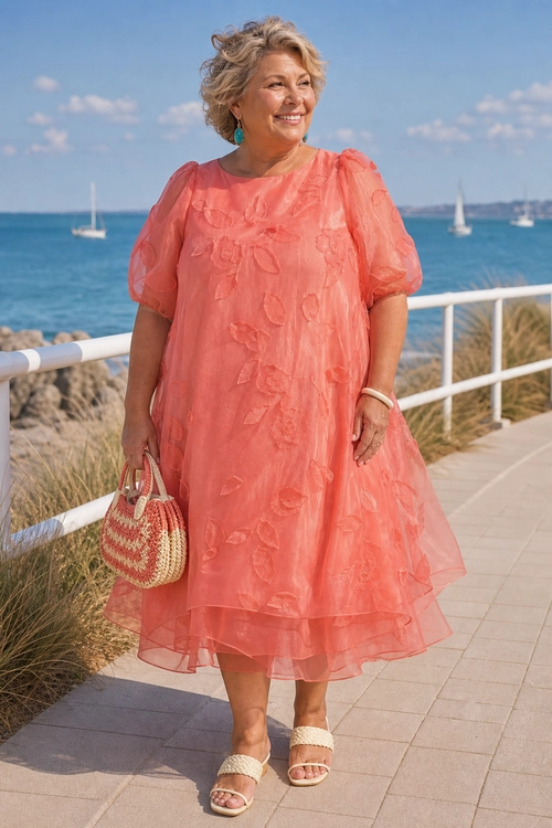 Coral Floral Organza Dress by the Sea A woman wears a coral floral organza dress with puff sleeves, woven handbag, turquoise earrings, and beige sandals beside the sea.