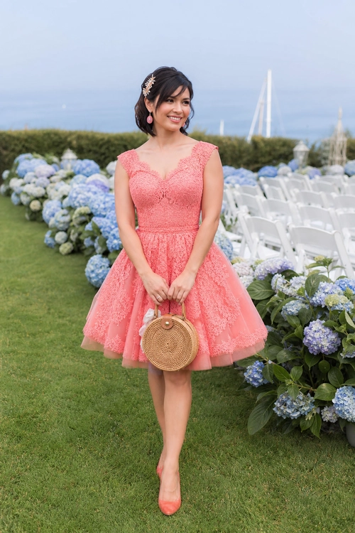Coral Lace Fit-and-Flare Dress Woman wearing a coral lace fit-and-flare dress with matching heels and a round woven handbag in a flower-filled garden.