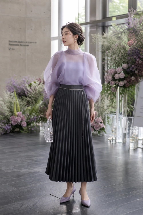 A woman wears a sheer lavender puff-sleeve blouse with a black pleated midi skirt, lavender pointed-toe heels, and a silver crystal clutch.