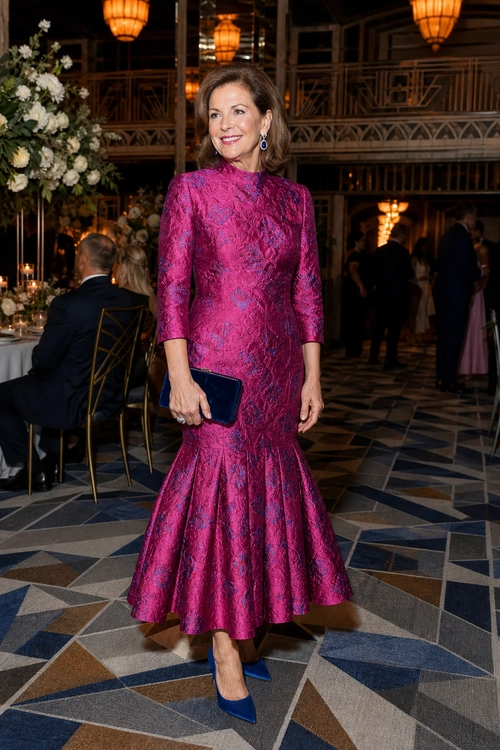 Magenta Floral Brocade Dress Woman wearing a long-sleeved magenta floral-patterned dress with a flared skirt, blue pointed heels, and a matching blue clutch.