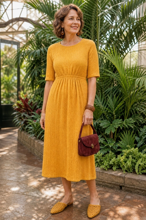 Mustard Textured Midi Dress Outfit A woman wears a mustard yellow textured midi dress with woven flats and carries a burgundy handbag in a leafy conservatory.