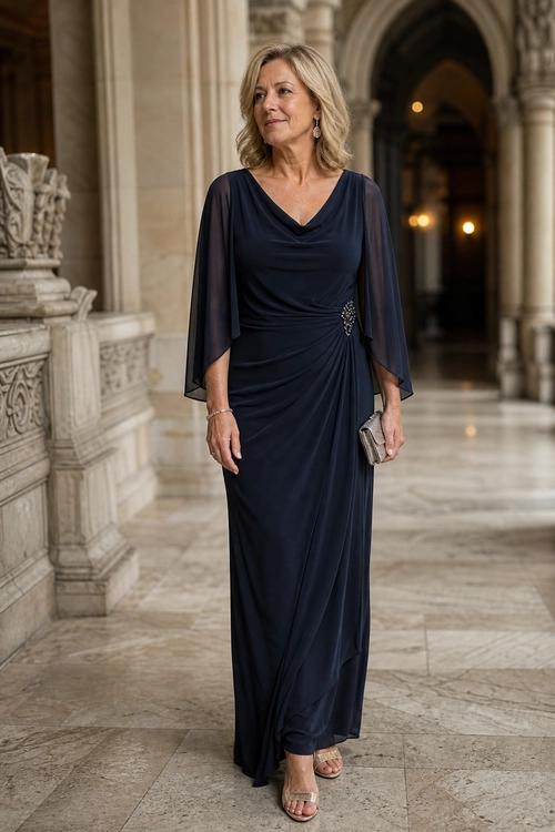 Navy Draped Evening Dress A woman wears a navy draped floor-length dress with sheer sleeves, a jeweled waist detail, a metallic clutch, and strappy sandals.