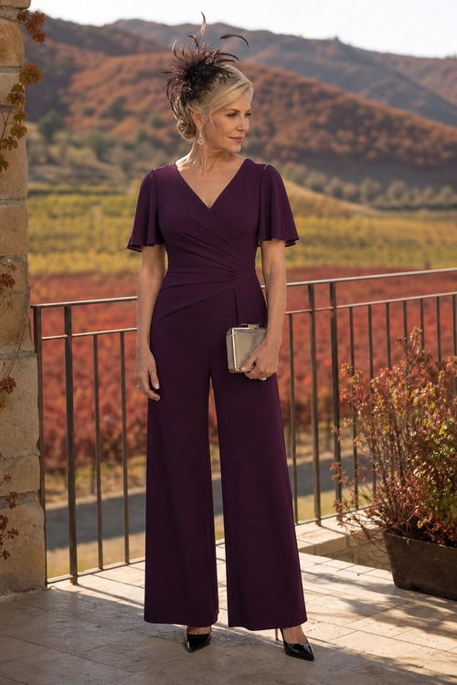 A woman wears a plum wide-leg jumpsuit with flutter sleeves, a feather fascinator, pointed-toe shoes, and a metallic clutch outdoors.