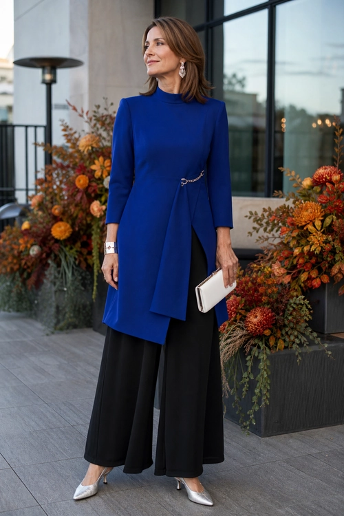 A woman wears a royal blue long-sleeve tunic with an asymmetric draped front over black wide-leg trousers, paired with pointed metallic heels and a white clutch.