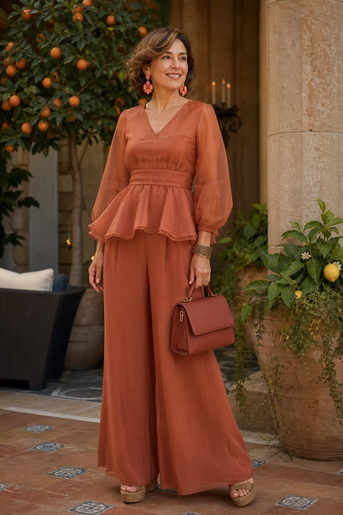 Woman wearing a rust-colored sheer-sleeve peplum top with matching wide-leg trousers, platform sandals, a structured handbag, earrings, and a bracelet.
