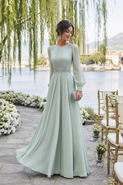 Woman wearing a flowing sage green long-sleeve gown with a beaded waist detail and a metallic clutch.