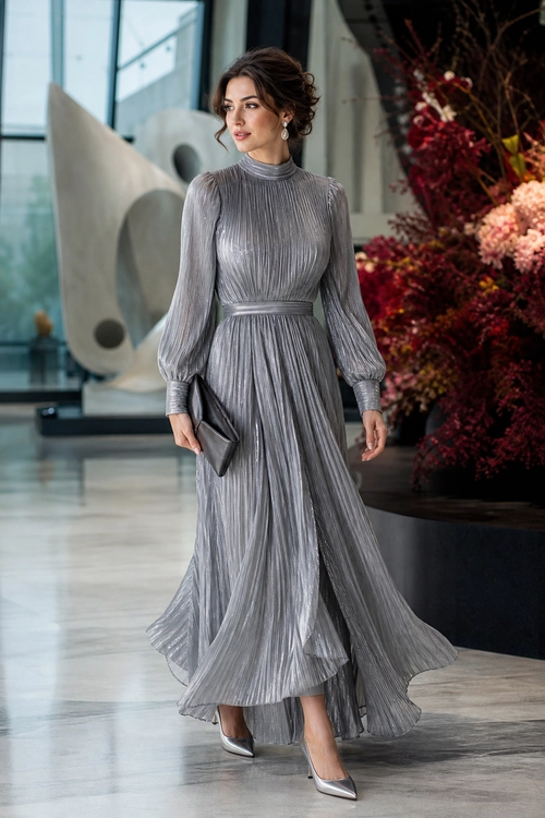 A woman wears a long silver-gray pleated dress with a high neckline, flowing skirt, dark clutch, drop earrings, and pointed metallic pumps.