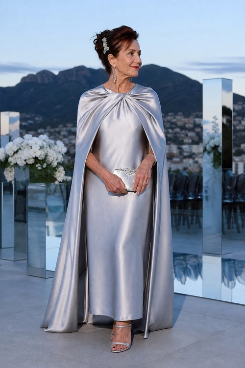 Silver Satin Cape Dress A woman wears a floor-length silver satin dress with a matching cape, metallic strappy sandals, dangling earrings, and a small embellished clutch.