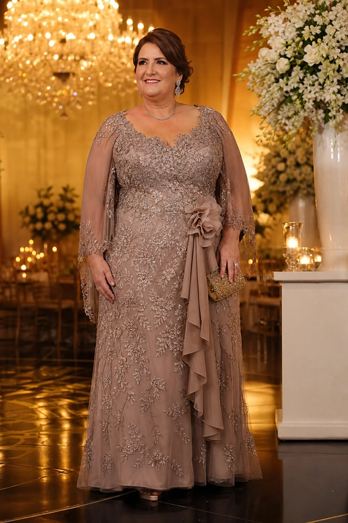 Taupe Embellished Evening Gown A woman wears a taupe floor-length gown with floral embellishment, sheer cape sleeves, a draped ruffle sash, and a gold clutch.