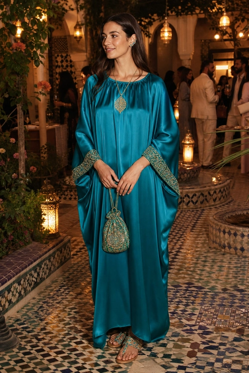 Woman wearing a flowing teal kaftan with embroidered cuffs, a statement pendant, embellished pouch, and matching flat sandals.