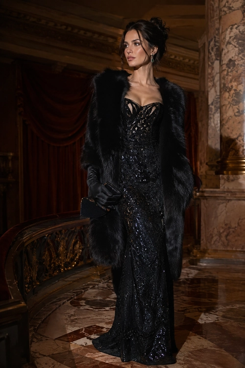 A woman wears a floor-length black sequin dress with a black fur coat, gloves, and clutch.